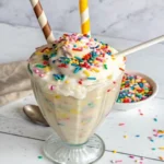 Vanilla protein shake in a tall glass topped with sprinkles, served with straw and spoon