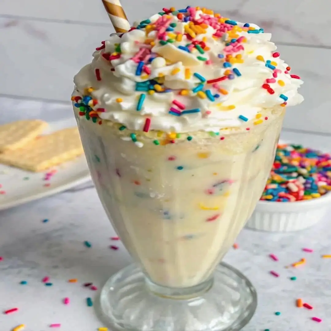 Thick birthday cake protein shake topped with whipped cream and colorful sprinkles in a tall glass