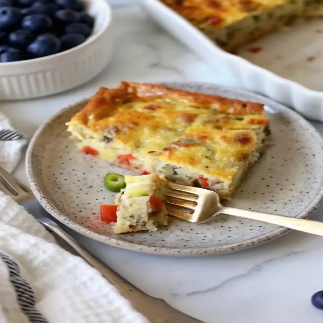 Slice of vegetable and sausage egg casserole on a ceramic plate with a fork and bowl of blueberries beside it