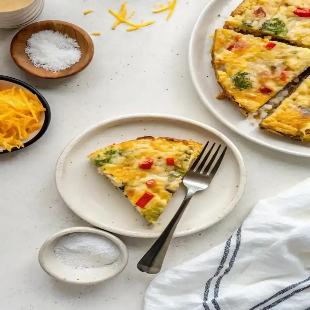 Slice of broccoli and cheese frittata on a white plate with two forks and extra shredded cheese nearby