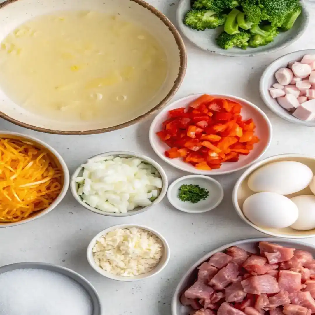 Bowls of eggs, egg whites, chopped vegetables, ham, cheese, and seasonings on a white counter