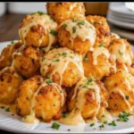 A tall stack of crispy, golden-brown fried potato balls drizzled with a rich cheese sauce and garnished with fresh parsley on a white plate.