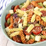 A light green bowl filled with cold summer pasta salad featuring spiral rotini, tomatoes, pepperoni, olives, and cheese on a wooden table.
