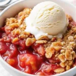 A white serving bowl filled with baked strawberry crumble and a scoop of vanilla ice cream melting into the warm fruit filling.