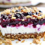 A slice of blueberry cheesecake bar showing distinct layers of crust, white cream filling, and dark berry topping with almonds and crumble.
