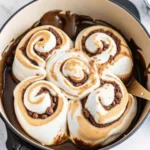 Close-up of cinnamon rolls in a skillet with melted chocolate sauce pooling between the rolls and toasted marshmallow topping.