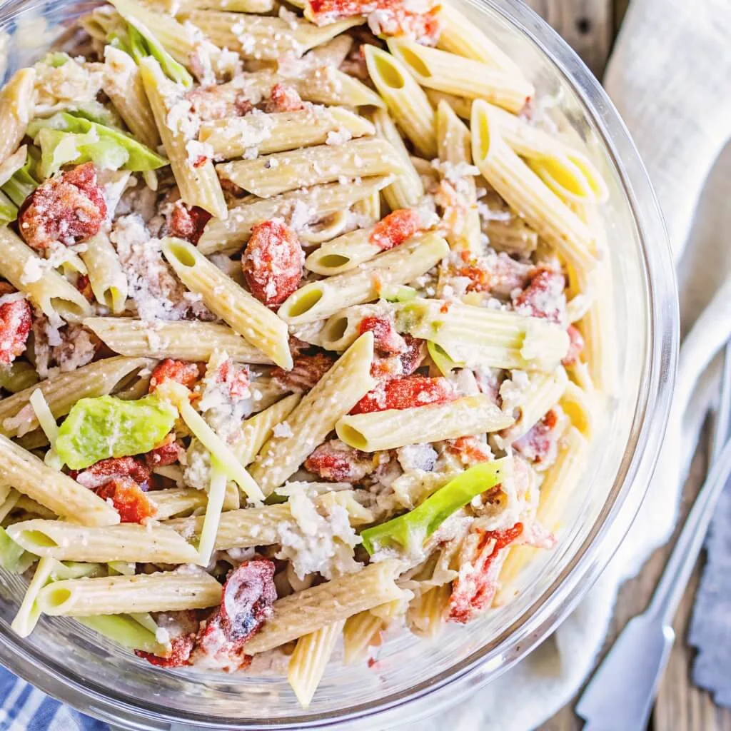 Close-up of penne pasta tossed with savory red tomatoes and fresh green herbs in a clear glass bowl.