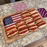 Rows of hot dogs with red toppings arranged on a wooden board that sits partially on top of an American flag.
