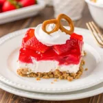 Close-up of a strawberry gelatin dessert slice with a pretzel on top, sitting on a white scalloped plate.