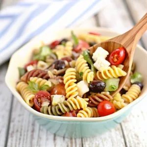 Close-up of a wooden spoon lifting Mediterranean pasta salad with spiral noodles, cherry tomatoes, black olives, cubed feta, and fresh basil.