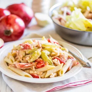 Close-up of a serving of penne pasta salad with tomatoes and greens on a white ceramic plate with a linen napkin.