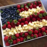 A festive fruit and cheese tray arranged like an American flag with fresh strawberries, blueberries, and cheese on a wooden table.