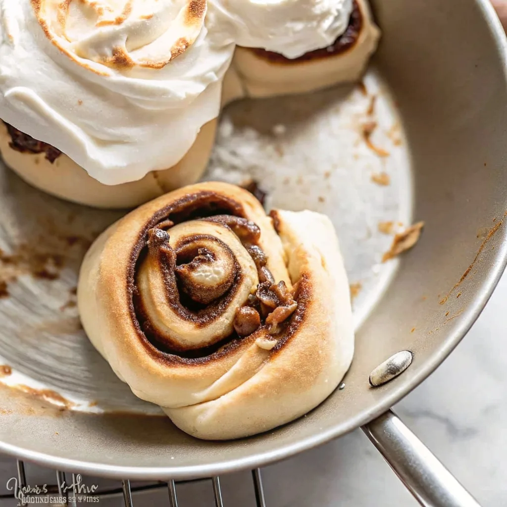 A single cinnamon roll sitting in a silver pan showing toasted edges, cinnamon sugar bits, and a large scoop of white frosting on the side.