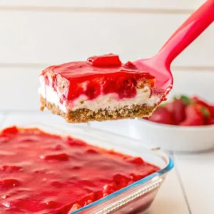 A square slice of layered strawberry dessert being held up on a pink spatula against a white background.