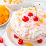 A high-angle shot of a large glass bowl containing ambrosia salad topped with cherries and oranges on a white marble surface.
