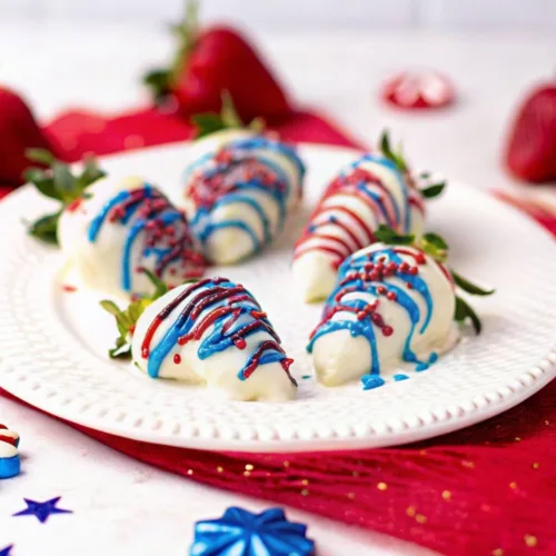 A white textured plate filled with strawberries dipped in white chocolate and drizzled with red and blue frosting, set against a red festive backdrop with star decorations.