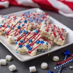 A white square plate filled with star-shaped rice krispie treats decorated with red frosting drizzles and blue star sprinkles, surrounded by scattered mini marshmallows.