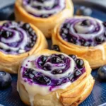 Close up of homemade blueberry sweet rolls with a swirling purple berry filling and dripping white cream cheese glaze on a blue plate.