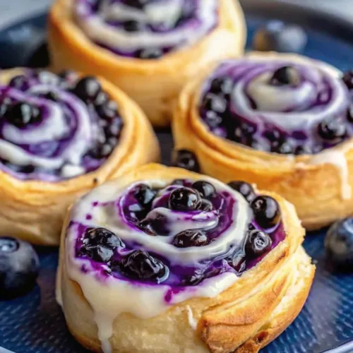 Blueberry Cheesecake Swirl Rolls 2 Close up of homemade blueberry sweet rolls with a swirling purple berry filling and dripping white cream cheese glaze on a blue plate.