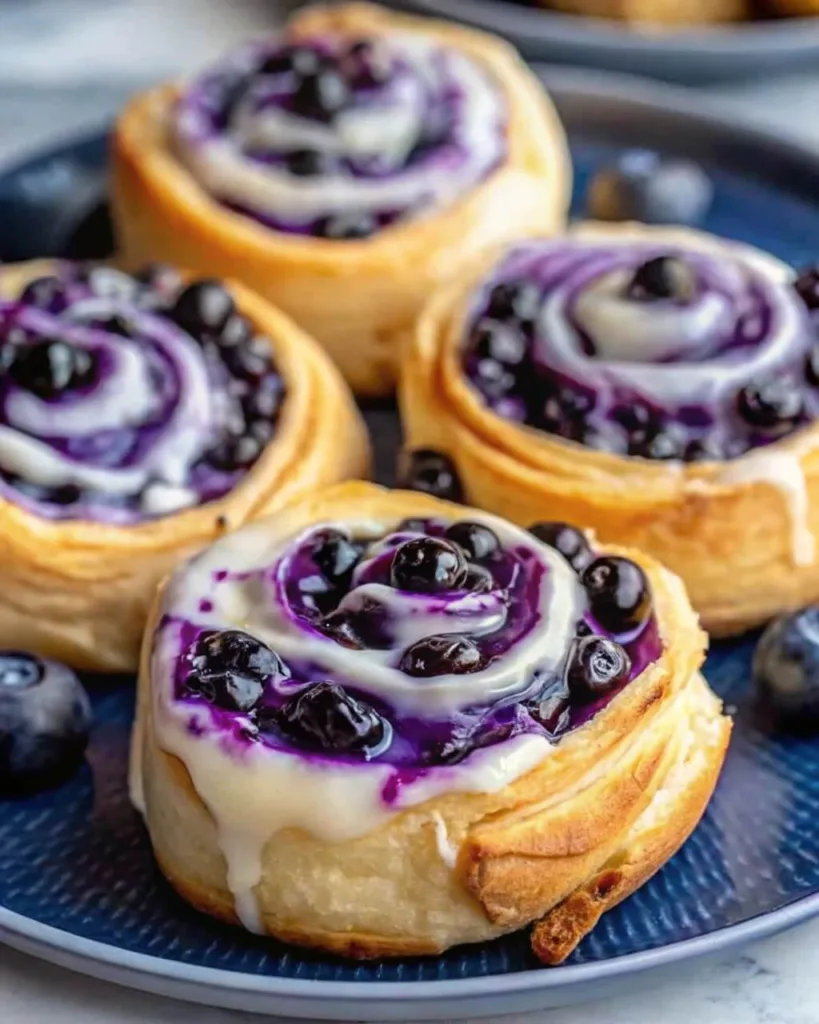 Close up of homemade blueberry sweet rolls with a swirling purple berry filling and dripping white cream cheese glaze on a blue plate.