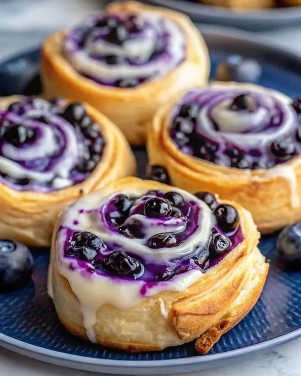 Blueberry Cheesecake Swirl Rolls 1 Close up of homemade blueberry sweet rolls with a swirling purple berry filling and dripping white cream cheese glaze on a blue plate.
