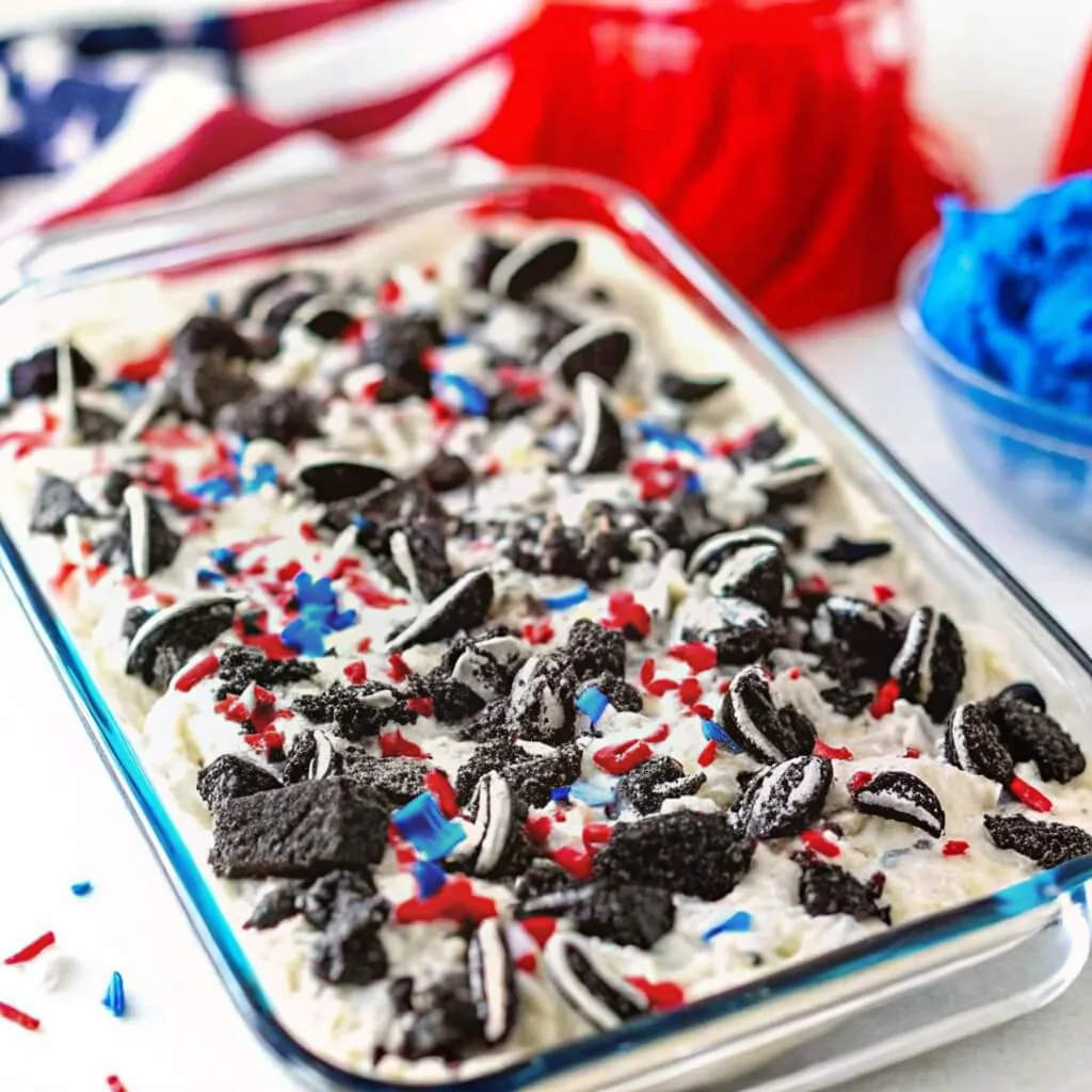 A glass baking dish filled with a patriotic Oreo poke cake topped with white frosting, chocolate cookie chunks, and red and blue star sprinkles.