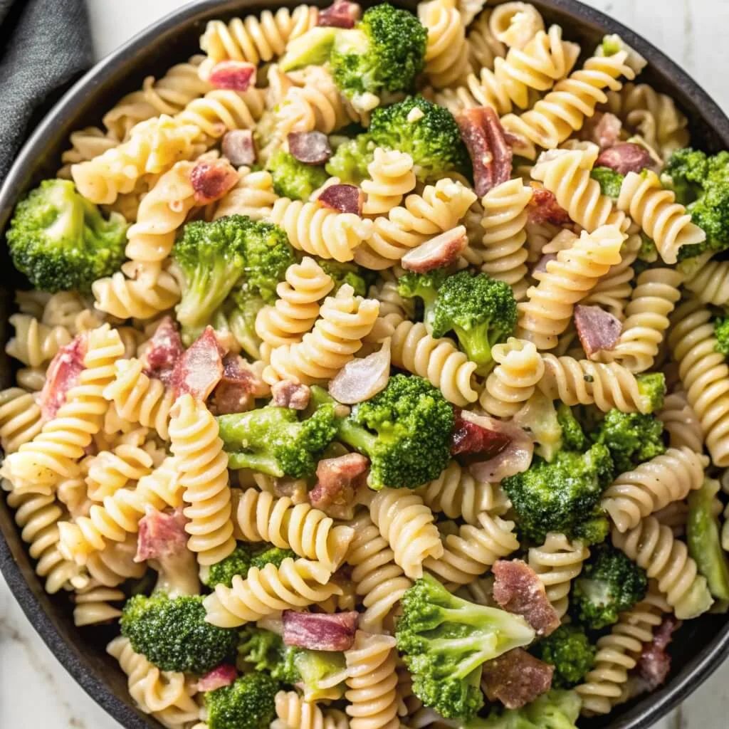 An overhead view of a black bowl filled with creamy rotini pasta, steamed broccoli florets, and crispy bacon pieces.