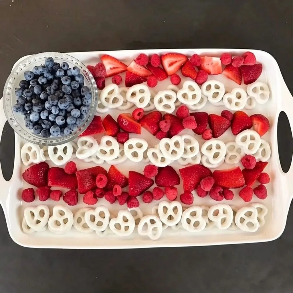 Festive Berry Flag Platter 2 An overhead view of a 4th of July charcuterie board shaped like an American flag with rows of strawberries, raspberries, yogurt covered pretzels, and a bowl of blueberries.