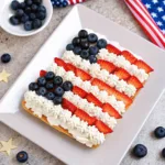 Two individual American flag fruit tarts served on a white marble pastry board with a bowl of blueberries in the background.