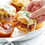 A hand picking up a cheesy meatball slider with melted mozzarella and marinara sauce on a white plate.
