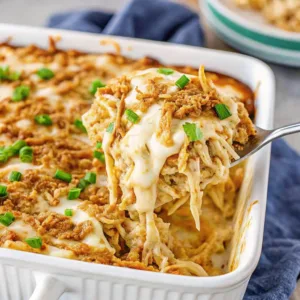A side-angle view of a spoon scooping a portion of shredded chicken casserole topped with melted cheese and crunchy fried onions.