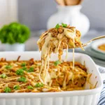 A serving spoon lifting a cheesy portion of French onion chicken from a white casserole dish, showing the bubbly melted cheese and herb garnish.