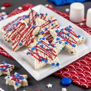 4th of July Rice Krispie Treats 3 A platter of star-shaped rice krispie treats with red frosting swirls and blue star sprinkles, displayed on a festive red and white patterned cloth.