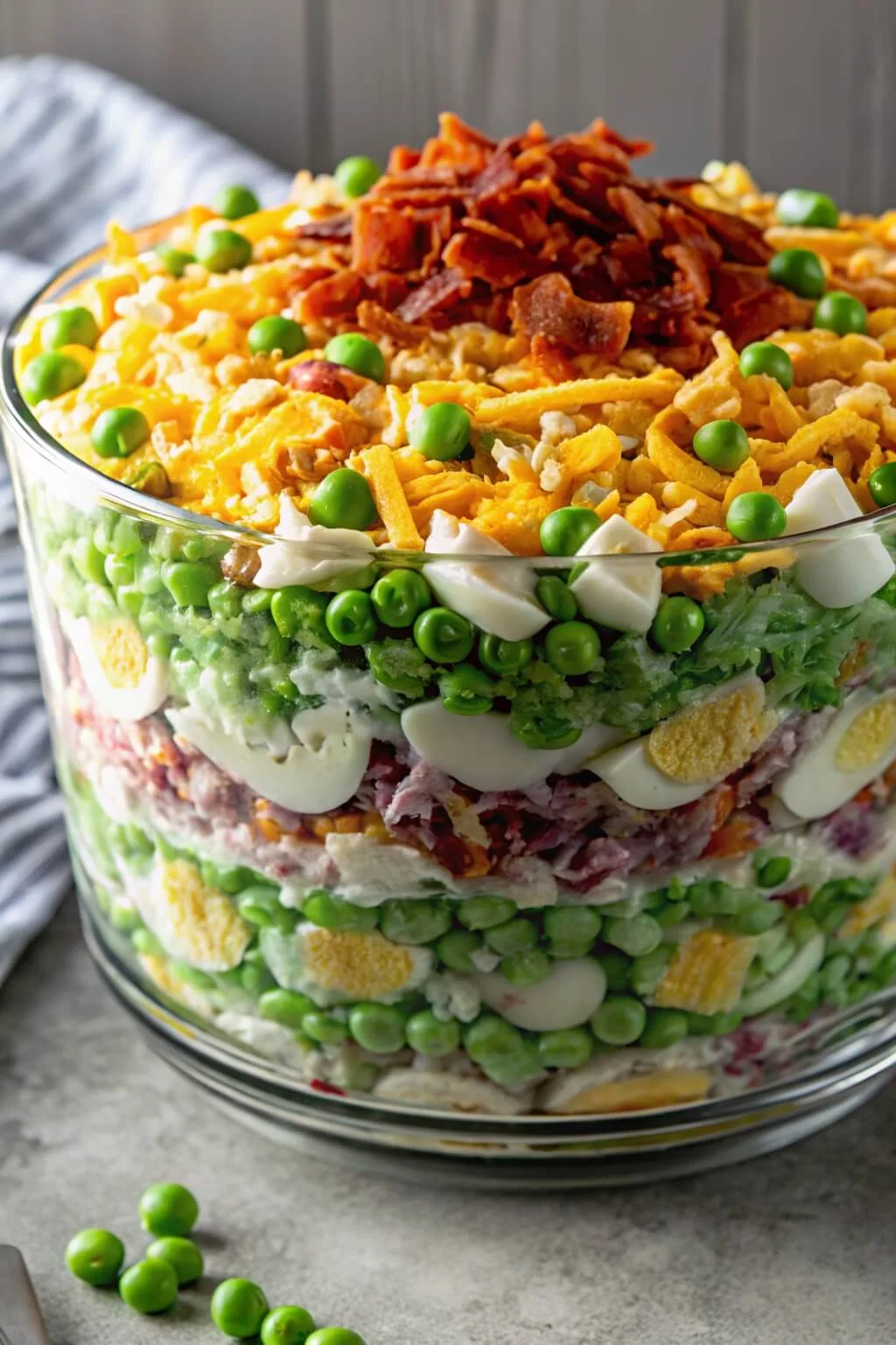 A side-angle view of a large glass bowl filled with a traditional layered overnight salad, showing distinct layers of fresh vegetables and eggs, topped with bacon.