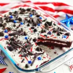 A close-up of a red velvet poke cake in a glass pan with a slice removed, showing the white cream filling and chocolate cookie topping.