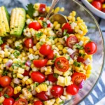 A large glass bowl showing the layers of a corn and tomato salad with red onion and cilantro.