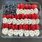 A patriotic american flag snack tray made with neat rows of fresh strawberries, yogurt pretzels, and blueberries in a square glass baking dish.