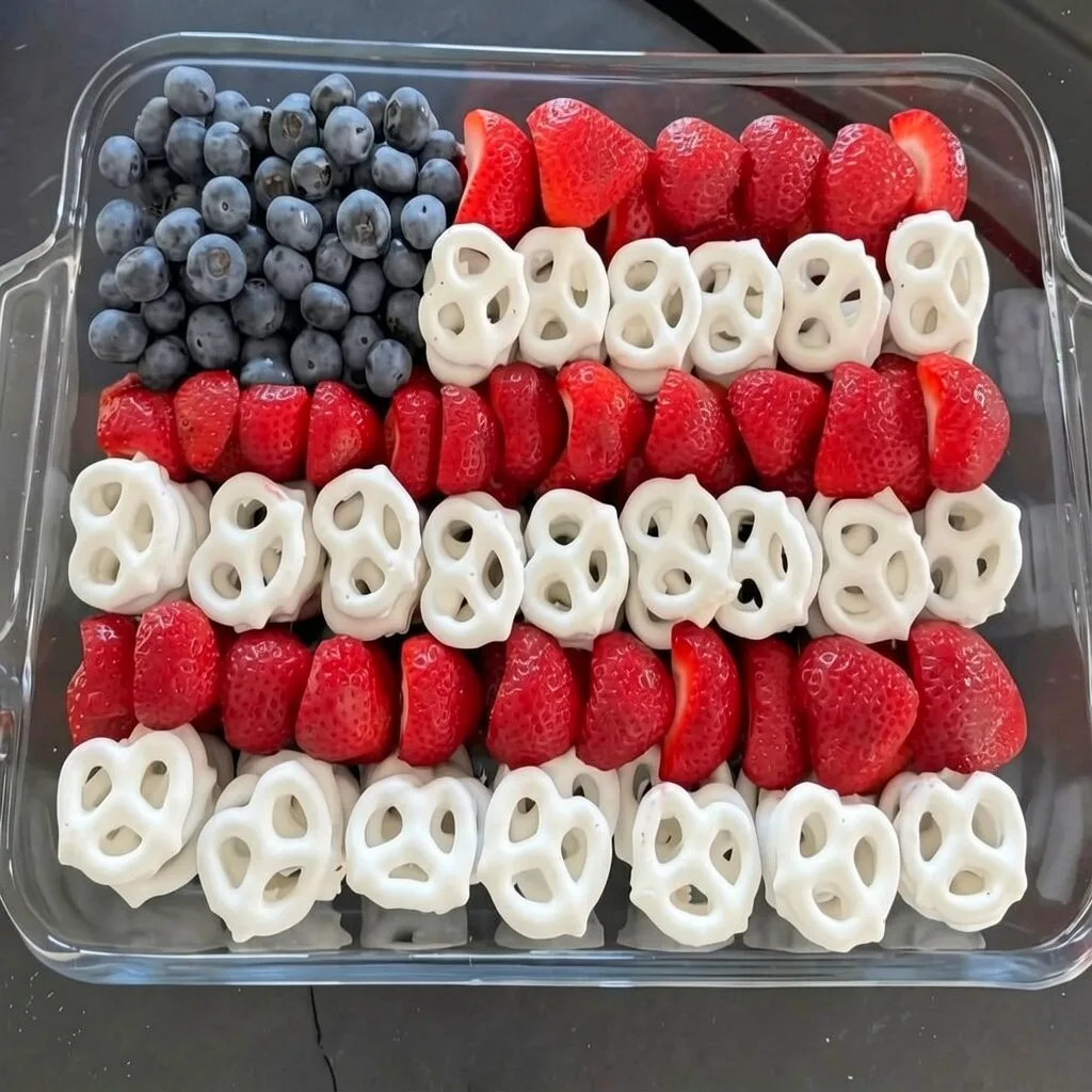 A patriotic american flag snack tray made with neat rows of fresh strawberries, yogurt pretzels, and blueberries in a square glass baking dish.