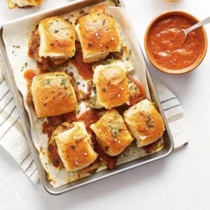 A baking sheet filled with baked meatball sliders topped with garlic herb butter and sesame seeds, served with marinara dipping sauce.
