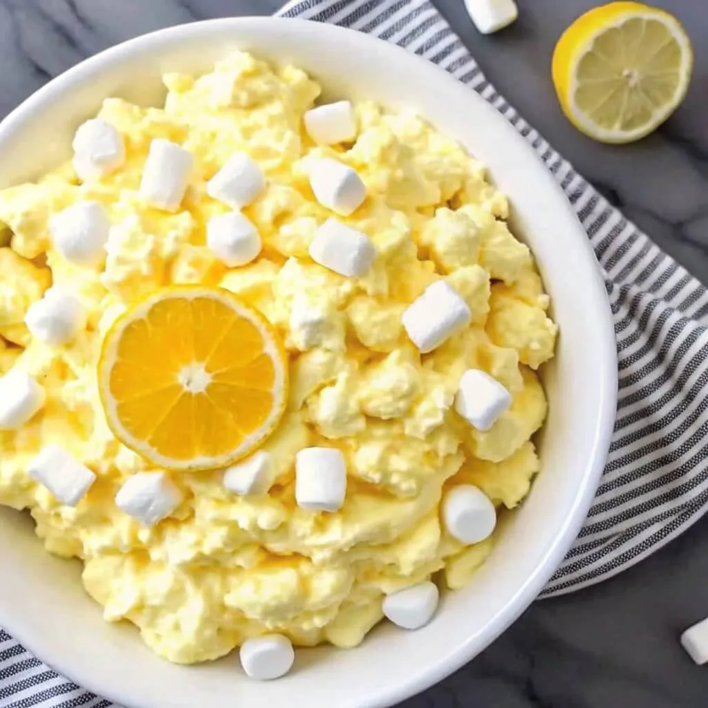 Classic Lemon Fluff Salad 3 Wide shot of a homemade lemon fluff dessert salad with marshmallows and a lemon slice garnish on a marble countertop.
