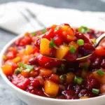 A gold spoon lifting a portion of cranberry mango salsa from a white bowl, highlighting the chunky texture and fresh herbs.