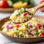 A wooden spoon lifting a serving of orzo salad with a tomato and cucumber garnish from a shallow brown ceramic bowl.