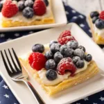 A square piece of puff pastry topped with white cream, fresh blueberries, and a red raspberry, dusted with sugar.
