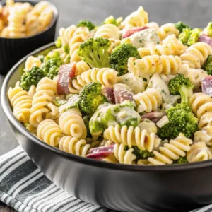A side-angle close-up of a loaded broccoli pasta salad in a black bowl sitting on a striped linen napkin.